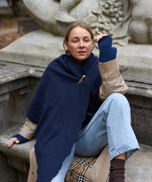 Eine Person, die einen „Homeland“-Schal von WALD Berlin x Helena Harfst trägt, an dem eine goldene Vogelbrosche befestigt ist, über einem beigen Trenchcoat und blauen Jeans, sitzt auf einem Steinsockel.