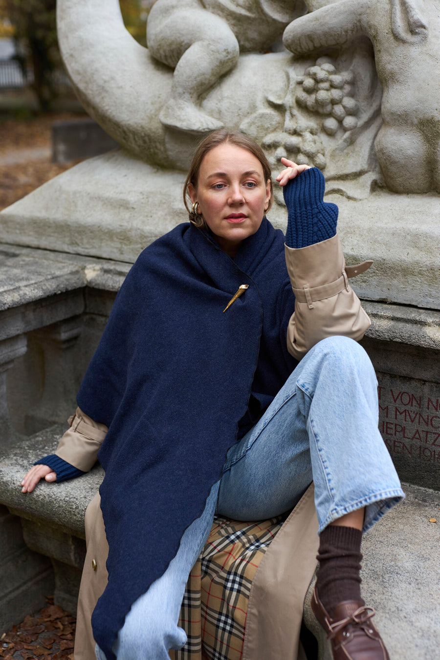 Eine Person, die einen „Homeland“-Schal von WALD Berlin x Helena Harfst trägt, an dem eine goldene Vogelbrosche befestigt ist, über einem beigen Trenchcoat und blauen Jeans, sitzt auf einem Steinsockel.