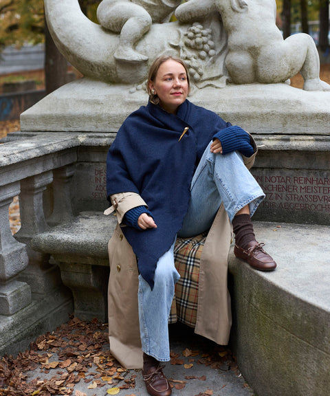 Eine Person, die einen marineblauen „Homeland“-Schal von WALD Berlin x Helena Harfst und einen hellbraunen Trenchcoat trägt, sitzt auf einer Steinbank vor einer Skulptur.