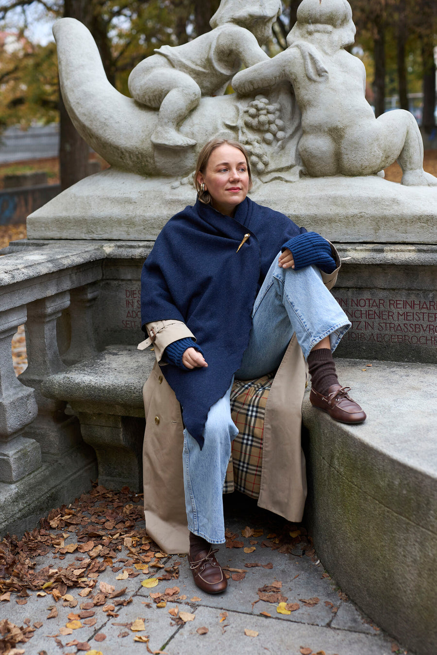Eine Person, die einen marineblauen „Homeland“-Schal von WALD Berlin x Helena Harfst und einen hellbraunen Trenchcoat trägt, sitzt auf einer Steinbank vor einer Skulptur.