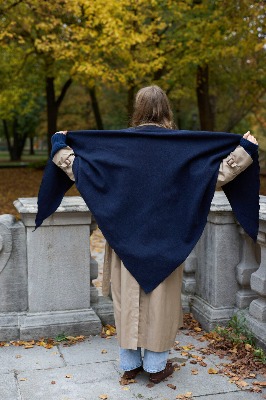 Eine Person, die von hinten zu sehen ist, trägt einen Trenchcoat und hält einen großen, marineblauen „WALD Berlin x Helena Harfst Homeland“-Schal offen hinter dem Rücken.