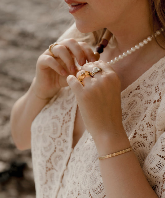 A woman wearing a lace blouse and an amber color bracelet is adjusting a pearl necklace. She has rings on her fingers, including the Isla Bonita Shining Star ring by WALD Berlin.