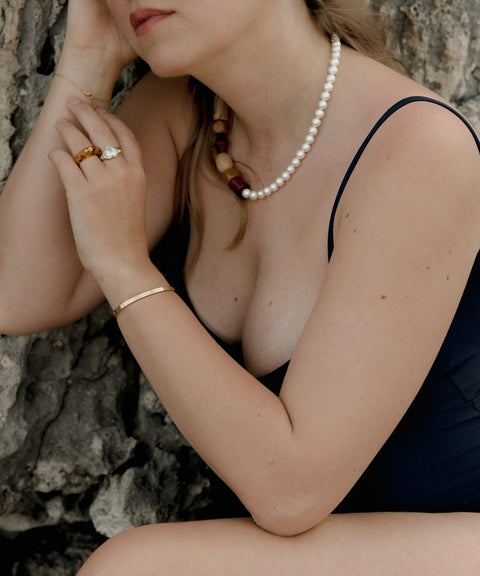 A person wearing a black swimsuit, pearl necklace, and gold jewelry, including the Isla Bonita Shining Star ring from WALD Berlin with an amber-colored gemstone, is seated against a textured rock surface, holding their hands near their face.