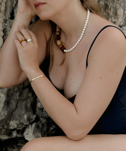 A person wearing a black swimsuit, pearl necklace, and gold jewelry, including the Isla Bonita Shining Star ring from WALD Berlin with an amber-colored gemstone, is seated against a textured rock surface, holding their hands near their face.