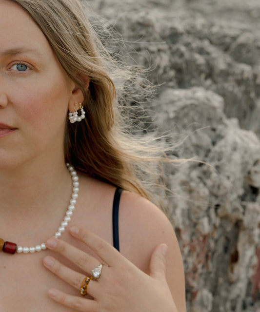 A woman with long blonde hair wears the Isla Bonita Pearl Hoops by WALD Berlin, along with a pearl necklace and rings, as she stands outdoors near rocks, her hand resting on her chest.
