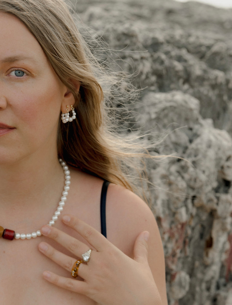 A woman with long blonde hair wears the Isla Bonita Pearl Hoops by WALD Berlin, along with a pearl necklace and rings, as she stands outdoors near rocks, her hand resting on her chest.