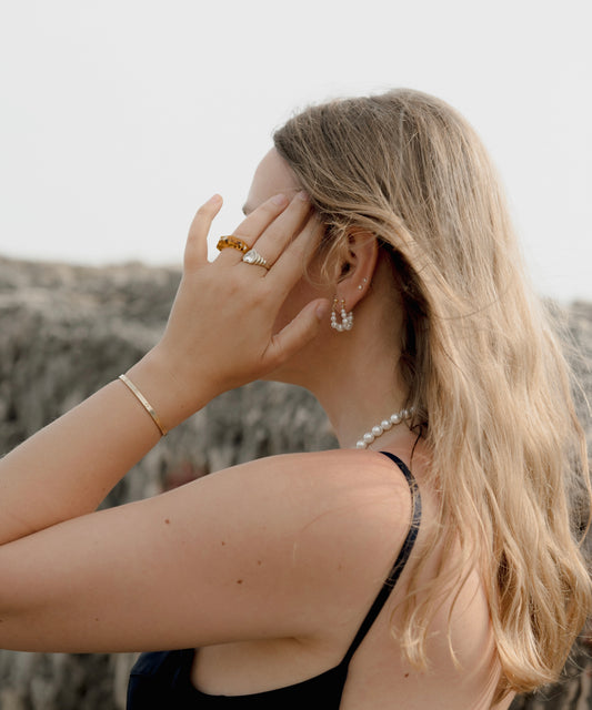 A woman with long blonde hair and a black top stands outdoors, facing away, touching her face. She wears WALD Berlin's Isla Bonita Pearl Hoops, a pearl necklace, and gold jewelry.