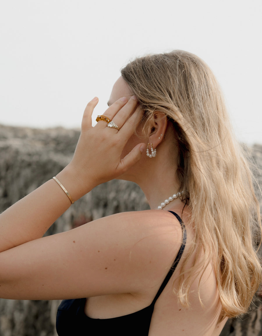 A woman with long blonde hair and a black top stands outdoors, facing away, touching her face. She wears WALD Berlin's Isla Bonita Pearl Hoops, a pearl necklace, and gold jewelry.
