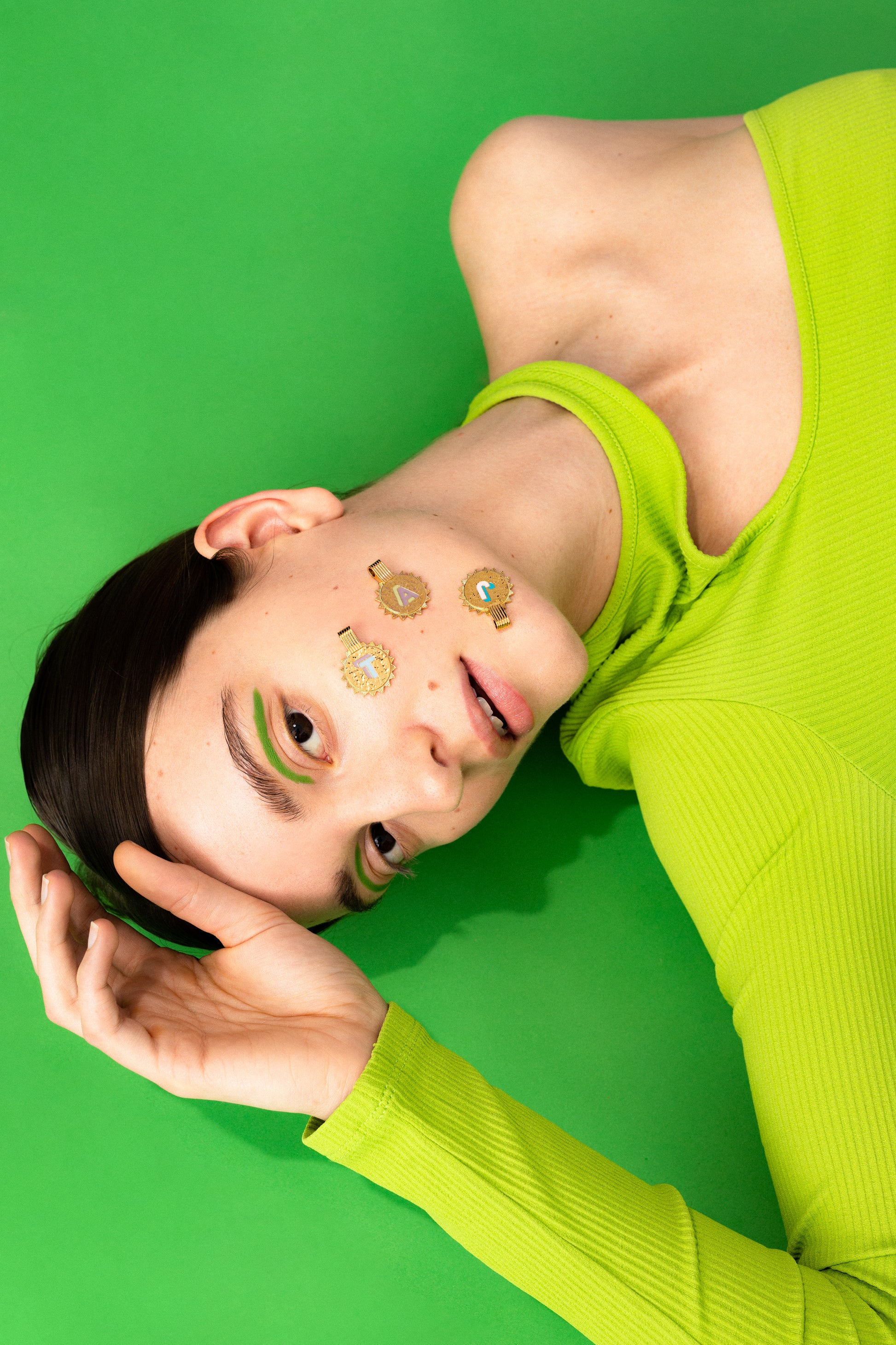 A person in a lime green top lies on a green surface with gold flower-shaped stickers, green makeup, and wears a WALD Berlin Sample saleSay My Name Gold Necklace of adjustable length around their neck.