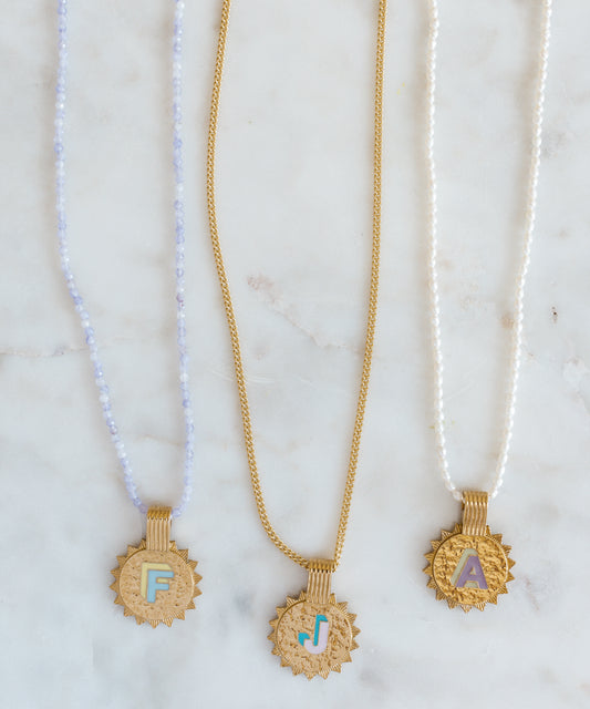 Three WALD Berlin Sample saleSay My Name Gold Necklaces, featuring gold-plated sunburst initials F, J, and A on blue beaded, gold, and pearl adjustable chains, are displayed on a white marble surface.