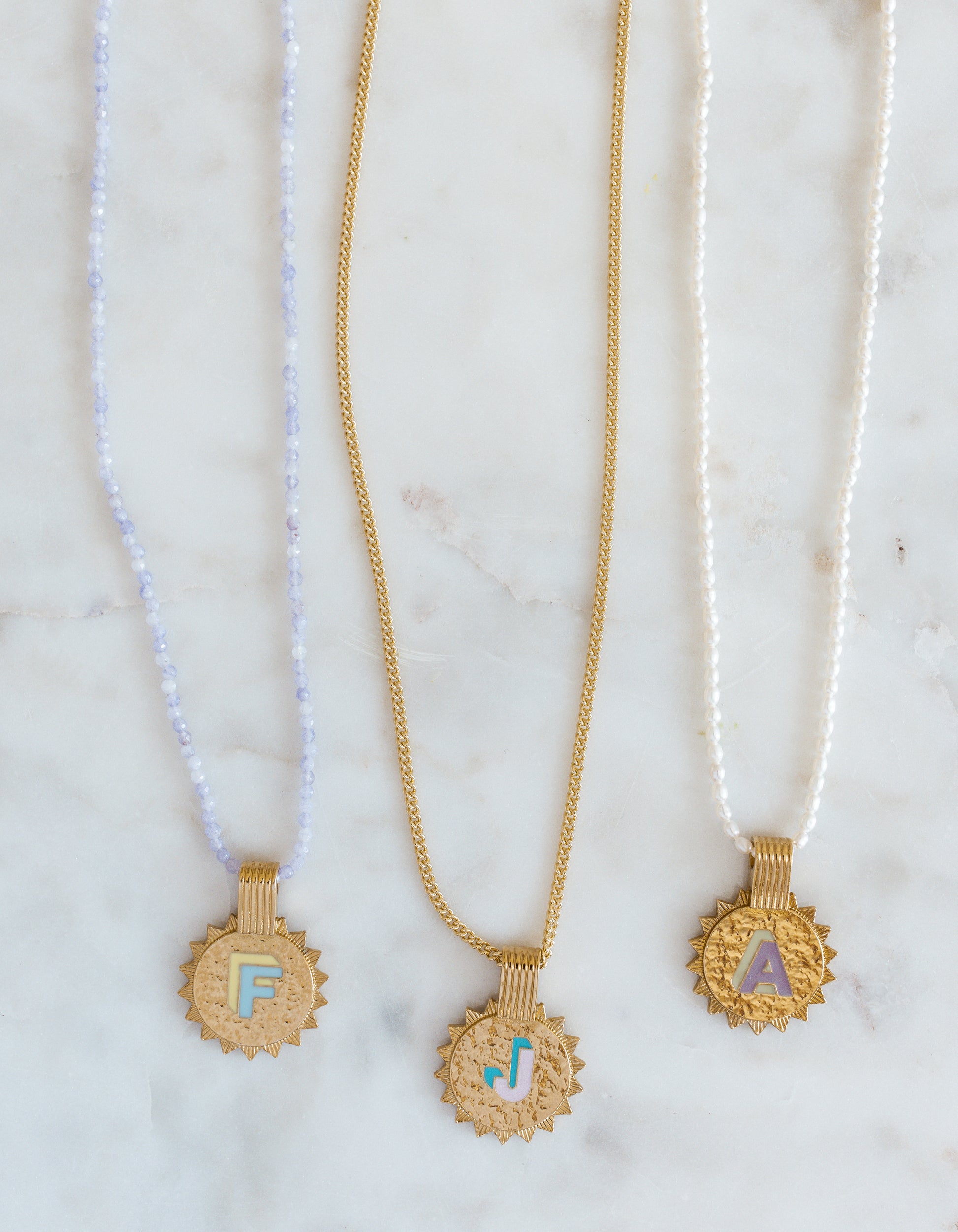 Three WALD Berlin Sample saleSay My Name Gold Necklaces, featuring gold-plated sunburst initials F, J, and A on blue beaded, gold, and pearl adjustable chains, are displayed on a white marble surface.
