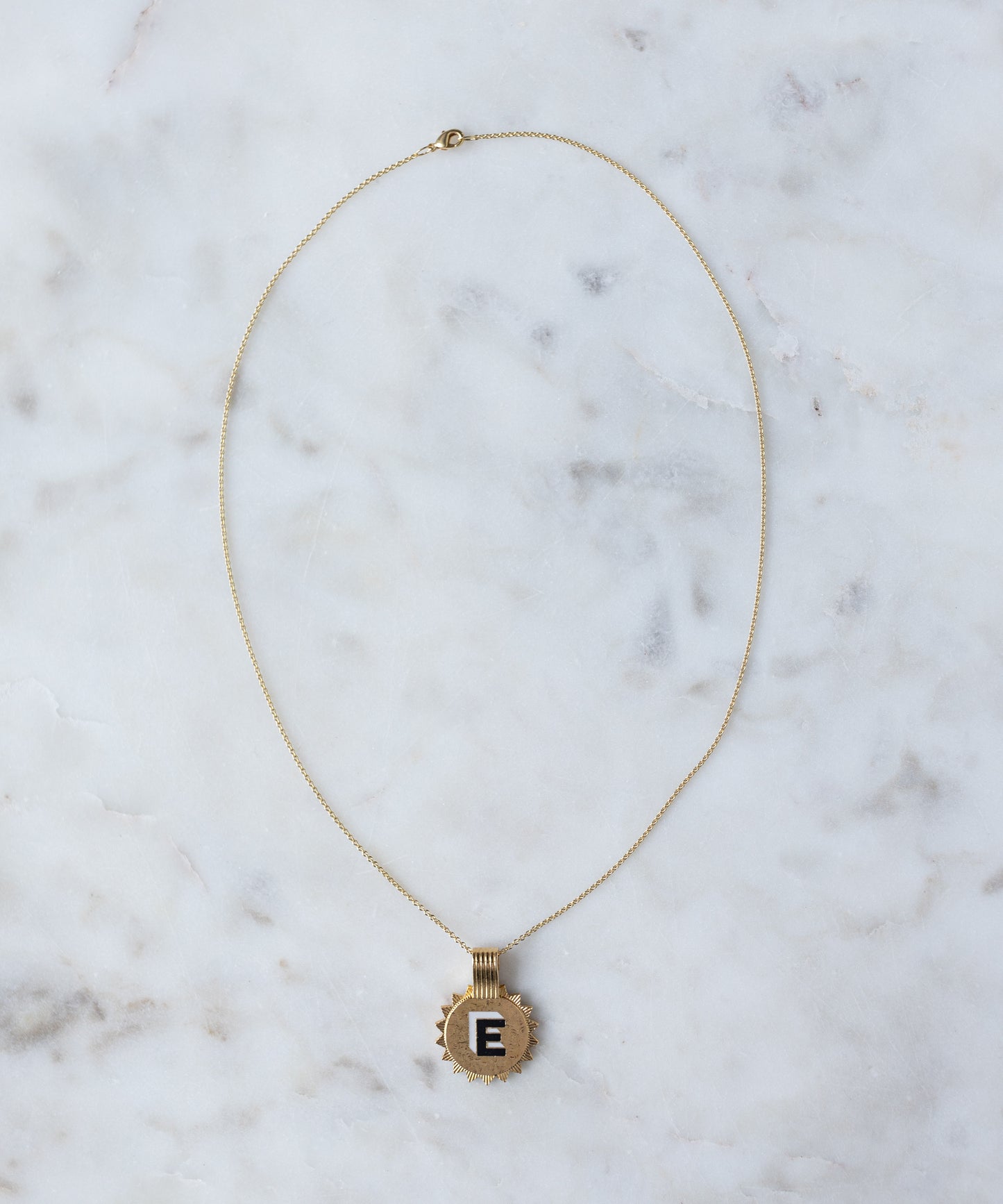The WALD Berlin Sample saleSay My Name Gold Necklace, a gold-plated initial necklace with a circular "E" pendant, rests on a white marble surface.