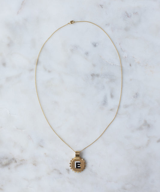 The WALD Berlin Sample saleSay My Name Gold Necklace, a gold-plated initial necklace with a circular "E" pendant, rests on a white marble surface.