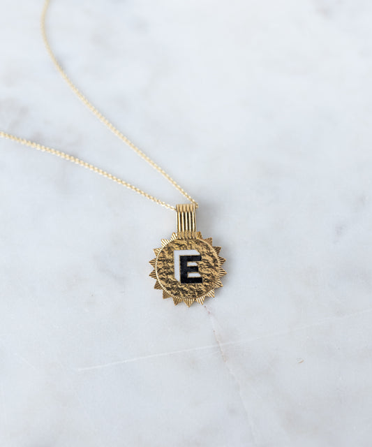 The WALD Berlin Sample Sale Say My Name Gold Necklace features a gold-plated round pendant with a black "E" initial, shown on light marble. Its adjustable length makes it ideal for layering or wearing solo.