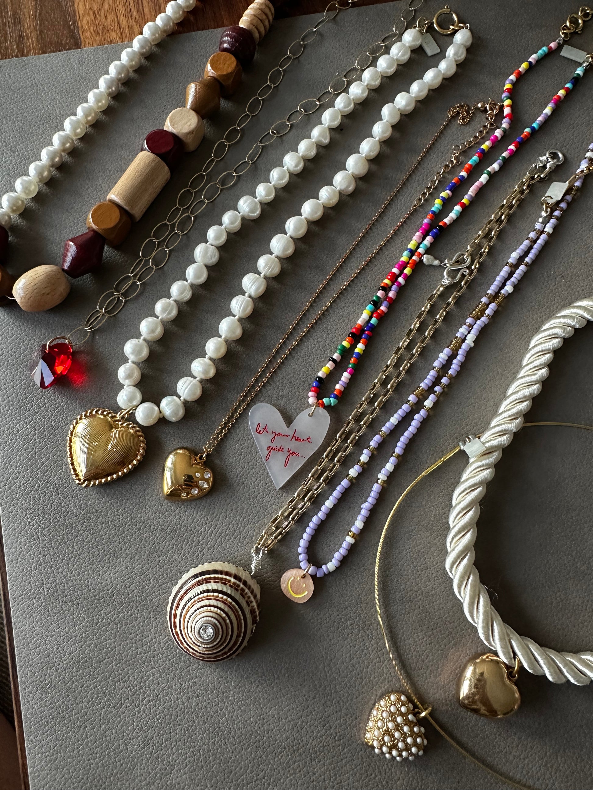A selection of necklaces with beads, pearls, vibrant patterns, gold-tone pendants, and a heart-shaped note displayed on grey—styled by Dana Roski for the signature WALD Berlin vibe. Featuring the surprise WALD Berlin Bag by WALD Berlin.
