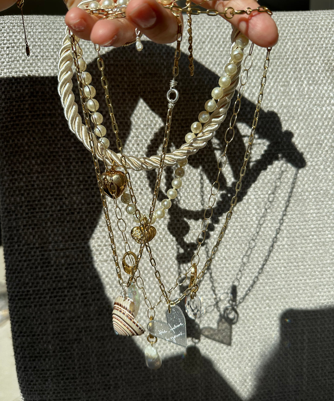 A hand holds several gold necklaces and a pearl choker from the surprise WALD Berlin Bag by WALD Berlin, featuring assorted pendants—shell, heart, and rectangular charm—set against a textured gray background.