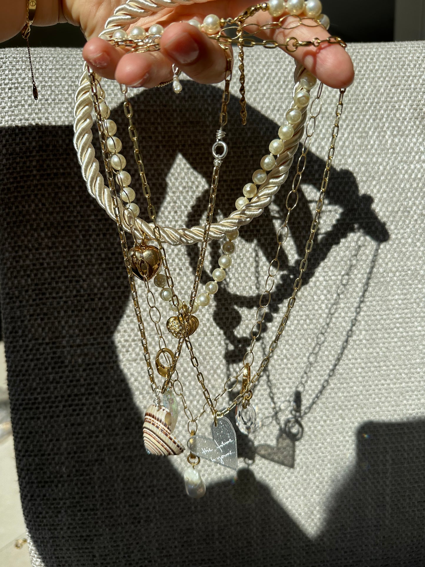 A hand holds several gold necklaces and a pearl choker from the surprise WALD Berlin Bag by WALD Berlin, featuring assorted pendants—shell, heart, and rectangular charm—set against a textured gray background.