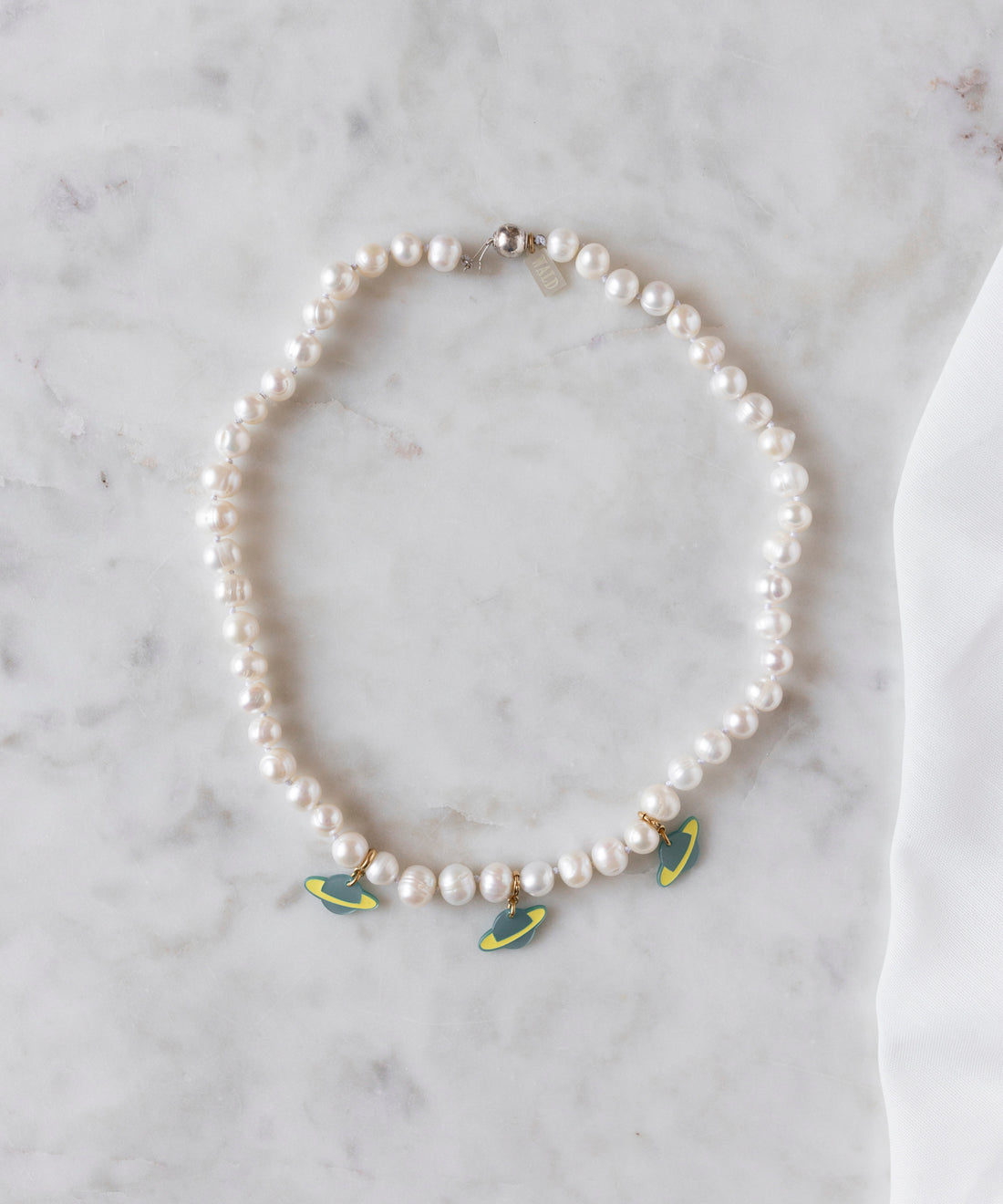 The WALD Berlin SAMPLE SALE From Another Mother Galactic Pearl Necklace, featuring sweetwater pearls and three green and yellow surfboard-shaped charms with 925 sterling silver accents, is displayed on a white marble surface.