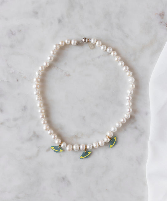 The WALD Berlin SAMPLE SALE From Another Mother Galactic Pearl Necklace, featuring sweetwater pearls and three green and yellow surfboard-shaped charms with 925 sterling silver accents, is displayed on a white marble surface.