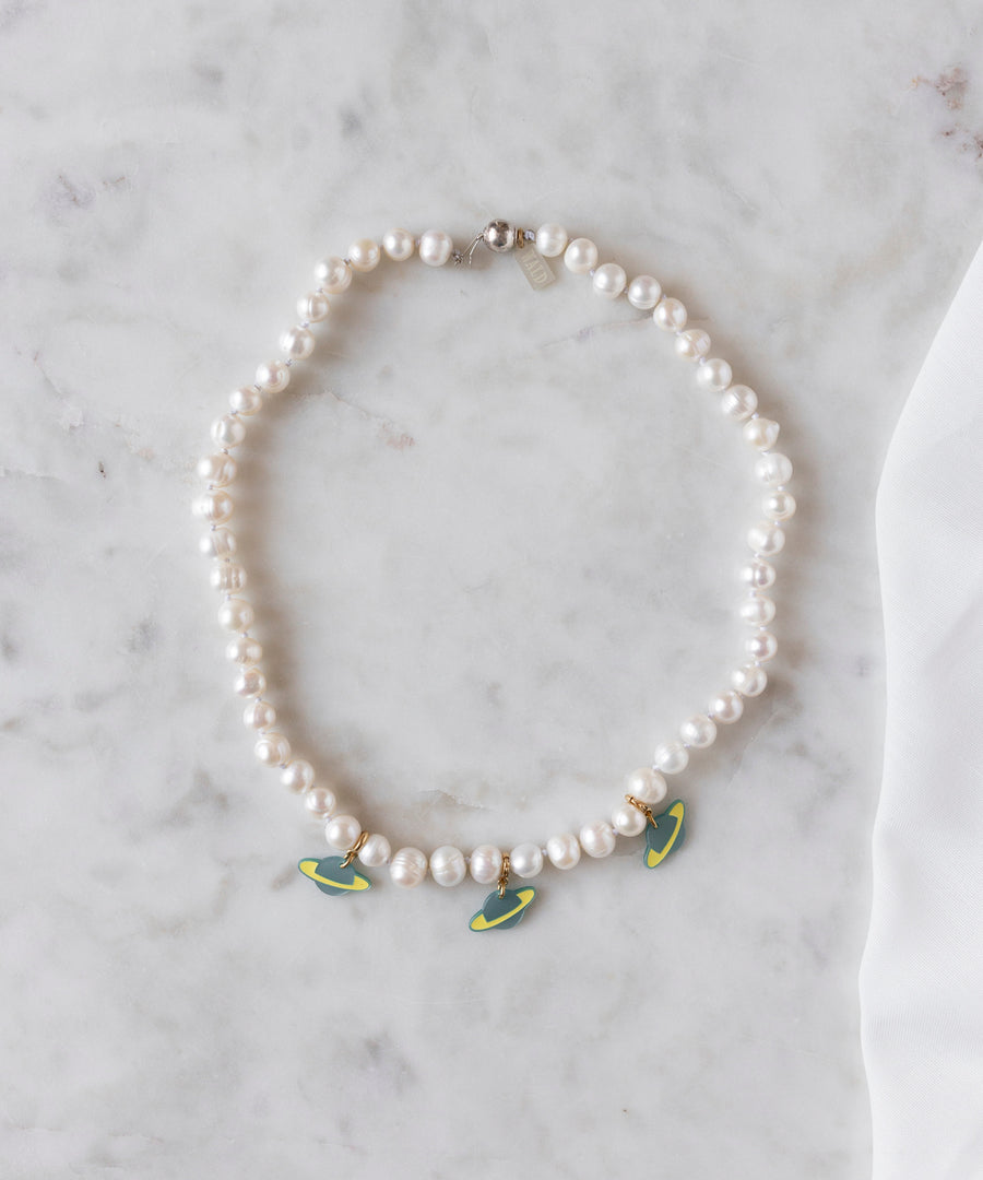 The WALD Berlin SAMPLE SALE From Another Mother Galactic Pearl Necklace, featuring sweetwater pearls and three green and yellow surfboard-shaped charms with 925 sterling silver accents, is displayed on a white marble surface.