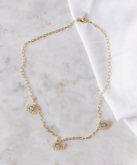 The SAMPLE SALE Daisy How High Necklace by WALD Berlin features an 18k gold-plated chain with three blue and gold sun-shaped charms, displayed on marble beside a white cloth—an ideal piece for everyday wear.