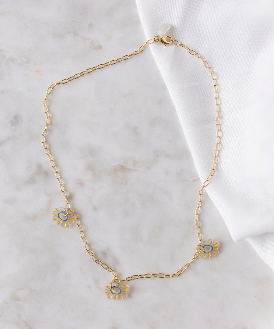 The SAMPLE SALE Daisy How High Necklace by WALD Berlin features an 18k gold-plated chain with three blue and gold sun-shaped charms, displayed on marble beside a white cloth—an ideal piece for everyday wear.