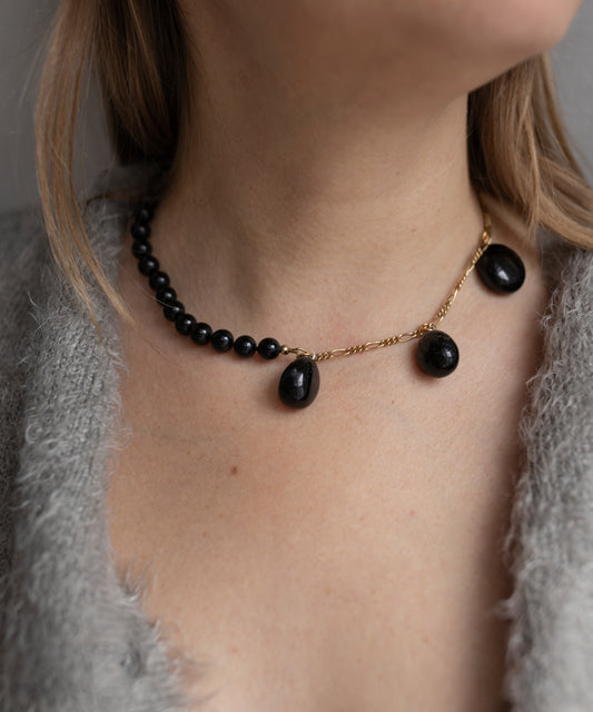 A person wearing the WALD Berlin Collection Noir Pas De Deux Black Drop Necklace, featuring black beads, an 18k gold plated chain, and three black pendants, paired with a gray fuzzy garment.