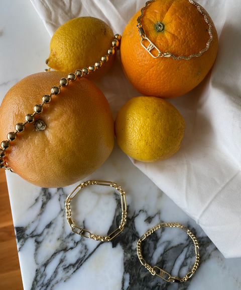 The Irina Gold Bracelets by WALD Berlin are elegantly draped over citrus fruits, including a grapefruit, orange, and lemons, on a marble surface with a white cloth.