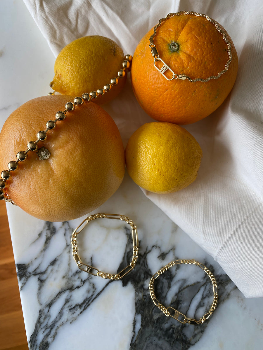 The Irina Gold Bracelets by WALD Berlin are elegantly draped over citrus fruits, including a grapefruit, orange, and lemons, on a marble surface with a white cloth.