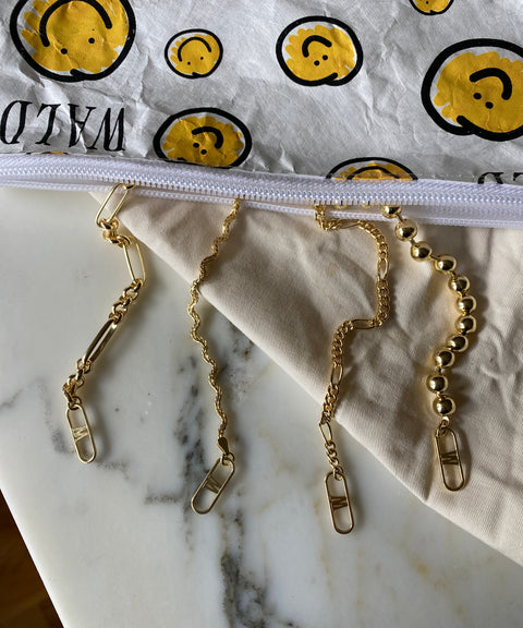 The Harry Necklace Gold by WALD Berlin, crafted from recycled gold and showcasing diverse link styles, is elegantly displayed on a marble surface. A white zipper pouch embellished with cheerful yellow smiley faces is partially visible.
