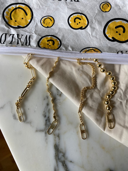 The Irina Necklace Gold by WALD Berlin, crafted from gold-plated brass and featuring diverse link styles, elegantly rests on a white marble surface. Nearby, packaging embellished with smiley faces showcases the artistry of German jewelry design.