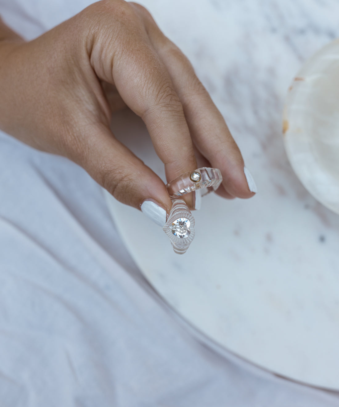 A hand with white-painted nails holds two transparent rings, including the WALD Berlin Lover Transparent Ring, adorned with a Swarovski stone, and another with a small gold accent, over a white marble surface.