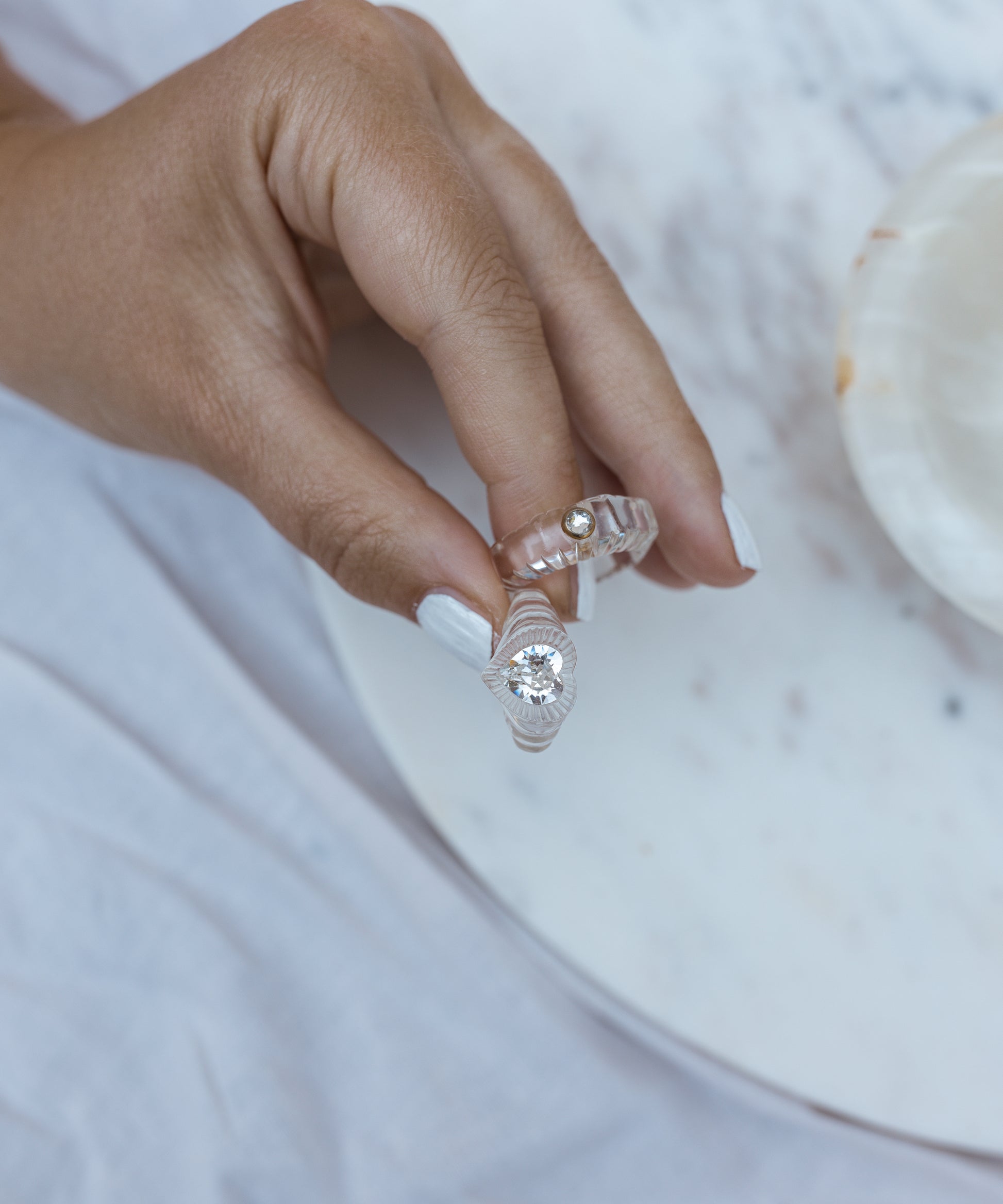A hand with manicured nails gracefully holds two rings against a light marble surface—one featuring a dazzling Swarovski stone and the other, the WALD Berlin Be My Lover Transparent Ring, captivating with its smaller yet equally mesmerizing clear gem.