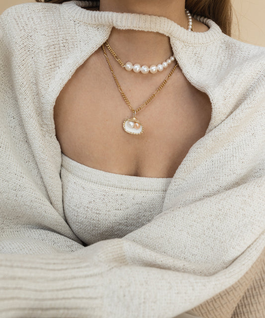 A person dons WALD Berlin's Pas De Deux Necklace, a handmade gold chain piece with a fair trade pearl pendant, paired with a cream knit sweater and top.