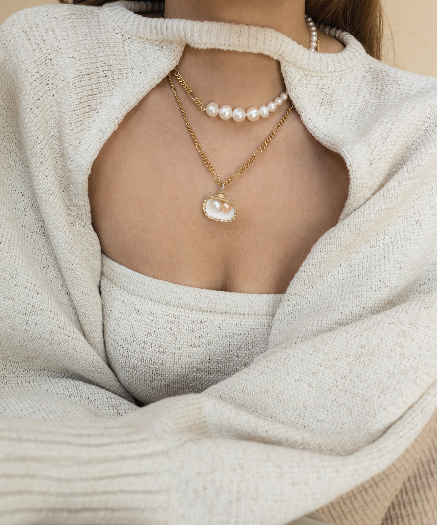 A person wears a cream sweater and top, accessorized with two necklaces: one delicate pearl strand and the WALD Berlin "Drop It Like It’s Hot Shell Necklace," featuring an exquisite gold-plated brass pendant.