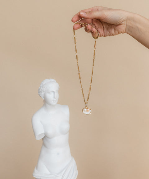 A hand displays the WALD Berlin "Drop It Like It's Hot" Shell Necklace in front of a small white bust sculpture of a woman, highlighting exquisite jewelry craftsmanship.