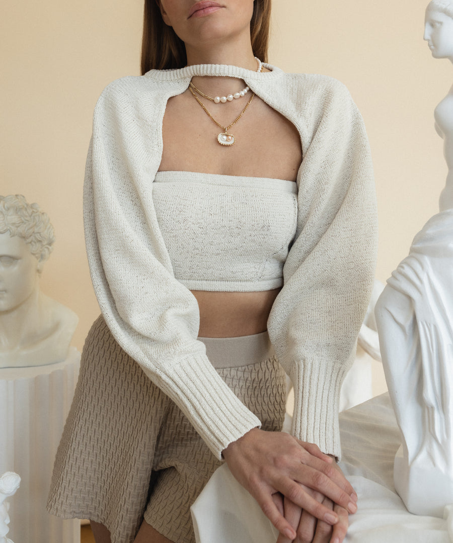 A person in a white knit top and beige shorts stands by sculptures in a neutral-toned room, their WALD Berlin Pas De Deux Necklace with a cultured pearl adding a touch of elegance.