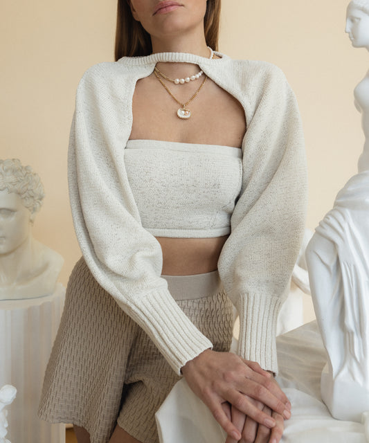 A person in a white knit shrug and tube top, adorned with WALD Berlin's Drop It Like It’s Hot Shell Necklace, stands near sculptures.
