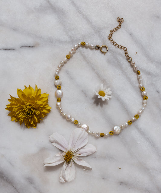 The Smilie Dude Pearl Necklace by WALD Berlin lies elegantly on marble, showcasing sweetwater pearls with smiley beads, surrounded by a vibrant yellow flower and delicate daisy-like blooms.