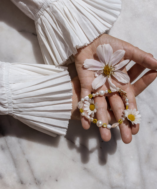 A hand with pleated, white sleeves holds a daisy, a cosmos flower, and WALD Berlin's Smilie Dude Pearl Bracelet on a marble surface.
