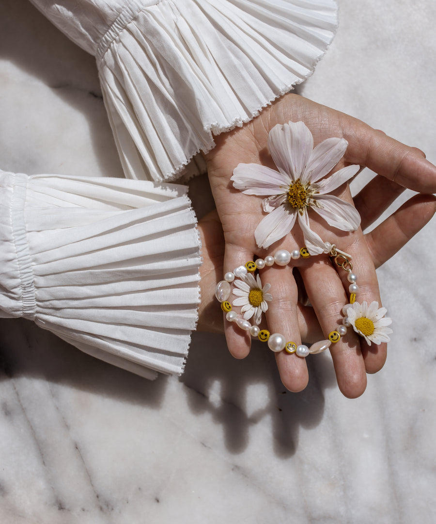 A hand with pleated, white sleeves holds a daisy, a cosmos flower, and WALD Berlin's Smilie Dude Pearl Bracelet on a marble surface.