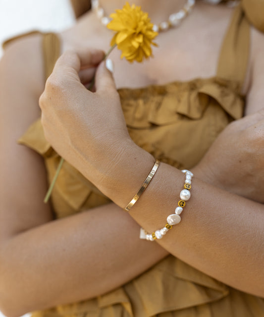 A person in a sleeveless dress holds a yellow flower, wearing WALD Berlin's Smilie Dude Pearl Bracelet with an extension on their wrist. This delightful jewelry features handmade touches, including exquisite sweetwater pearls.