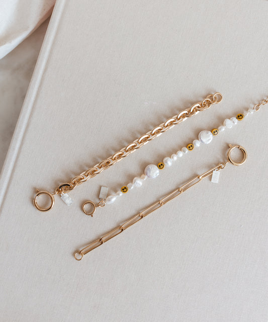 Three gold bracelets rest on a beige surface: an 18k gold-plated brass chain design, the WALD Berlin Smilie Dude Pearl Bracelet adorned with sweetwater pearls and smiley faces, and another featuring rectangular links.