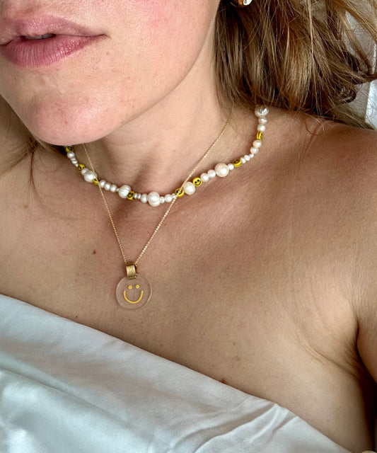 A close-up image shows a person wearing two necklaces. One necklace features pearls and smiley face beads, while the other is WALD Berlin's Smilie Dude Medaillon Gold Necklace, an 18k gold-plated piece with a transparent pendant made in Germany. The person is wrapped in a white sheet.