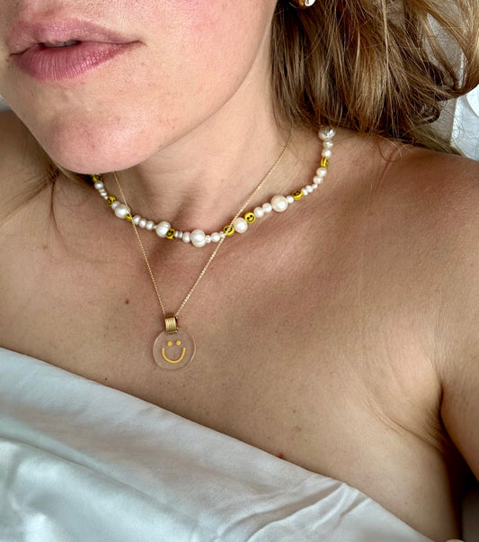 A close-up image shows a person wearing two necklaces. One necklace features pearls and smiley face beads, while the other is WALD Berlin's Smilie Dude Medaillon Gold Necklace, an 18k gold-plated piece with a transparent pendant made in Germany. The person is wrapped in a white sheet.