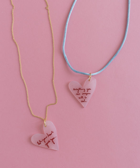 The "Let's be Happy Gold Necklace" by WALD Berlin features two heart-shaped pendants with inspirational text on an 18k gold-plated chain and blue cord, all against a pink background, inspiring positivity with every glance.