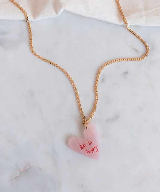 The WALD Berlin "Let's be happy" gold necklace features an 18k gold-plated chain with a pink heart-shaped hand-painted pendant inscribed with "don't be happy," stylishly displayed on a marble surface.
