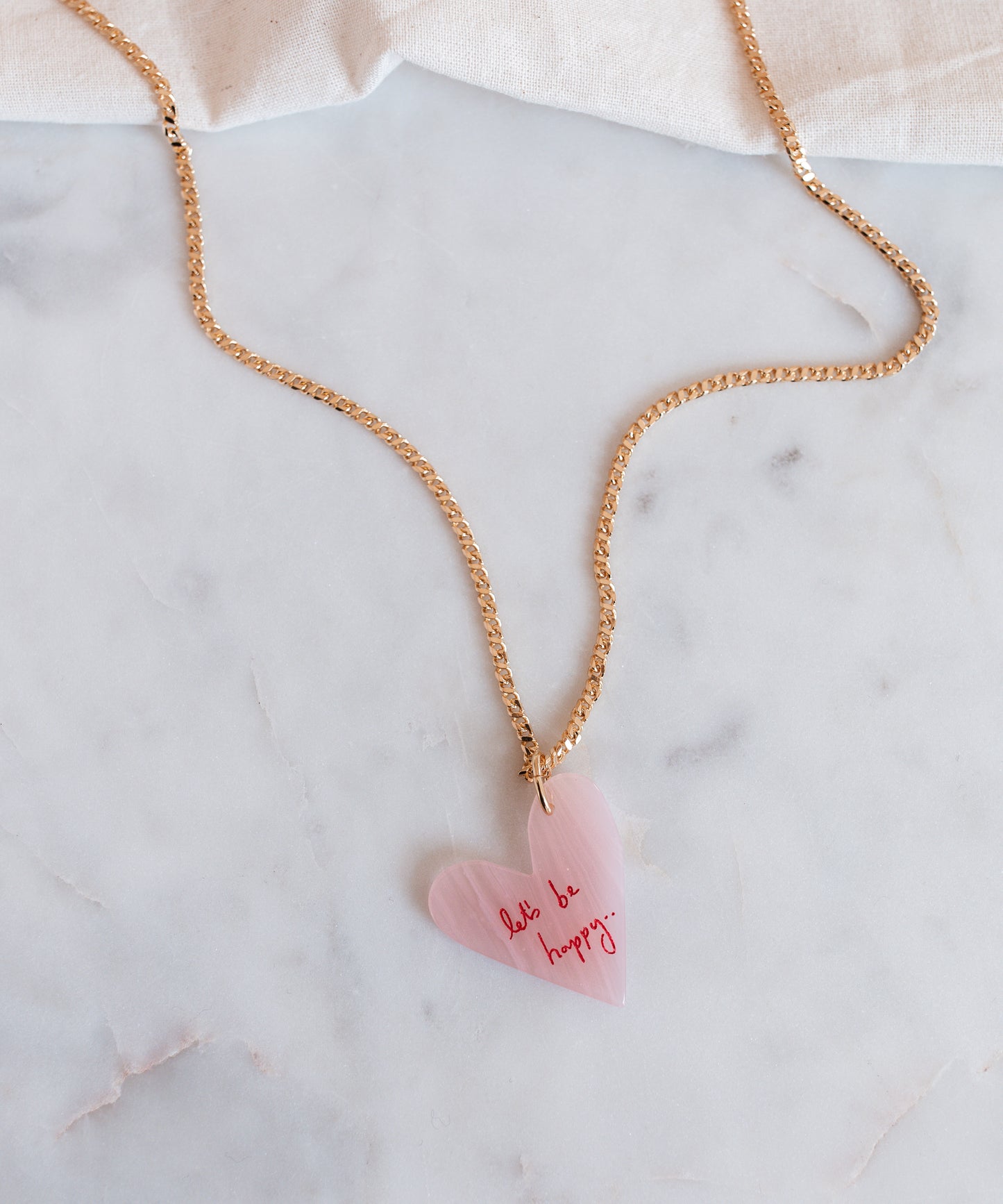 The WALD Berlin "Let's be happy" gold necklace features an 18k gold-plated chain with a pink heart-shaped hand-painted pendant inscribed with "don't be happy," stylishly displayed on a marble surface.