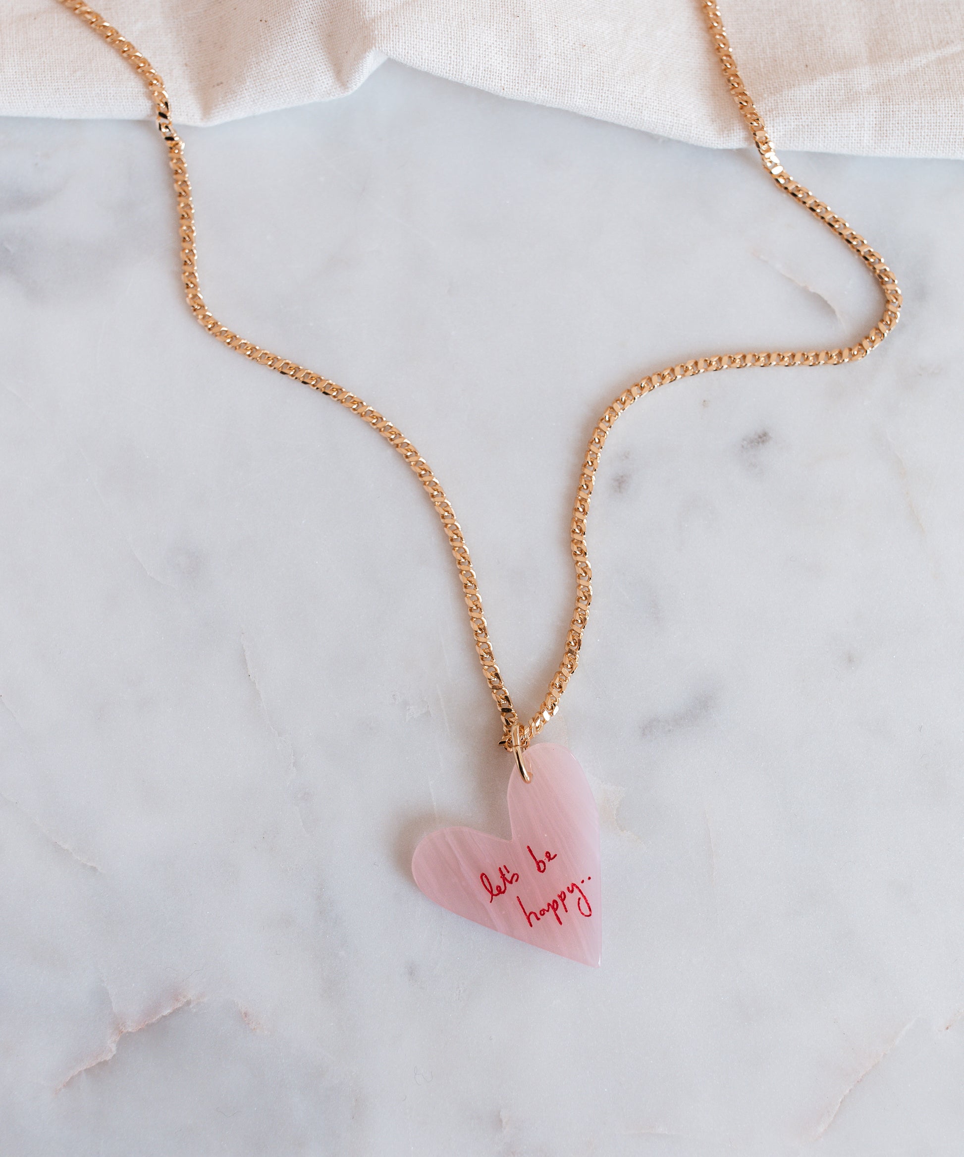 The WALD Berlin "Let's be happy" gold necklace features an 18k gold-plated chain with a pink heart-shaped hand-painted pendant inscribed with "don't be happy," stylishly displayed on a marble surface.