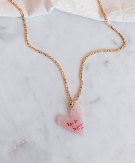 The WALD Berlin "Let's be happy" gold necklace features an 18k gold-plated chain with a pink heart-shaped hand-painted pendant inscribed with "don't be happy," stylishly displayed on a marble surface.